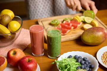 Fresh fruit being sliced beside two glasses of pink and green smoothies on wooden table. Concept of smoothie recipe blogs, nutrition lessons, healthy cooking tutorials, wellness marketing.