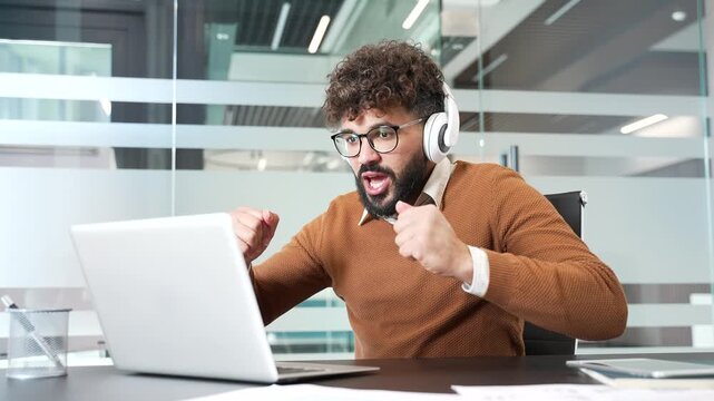 Happy excited businessman in headphones watching an online sports match using laptop sitting at workplace in business office. joyful entrepreneur cheering for bids at auction, celebrating success
