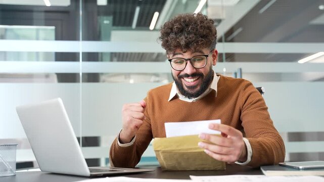 Joyful surprised businessman reading letter with great news sitting at workplace in glass business office. Smiling male professional is celebrating success, happy to receive positive good notification