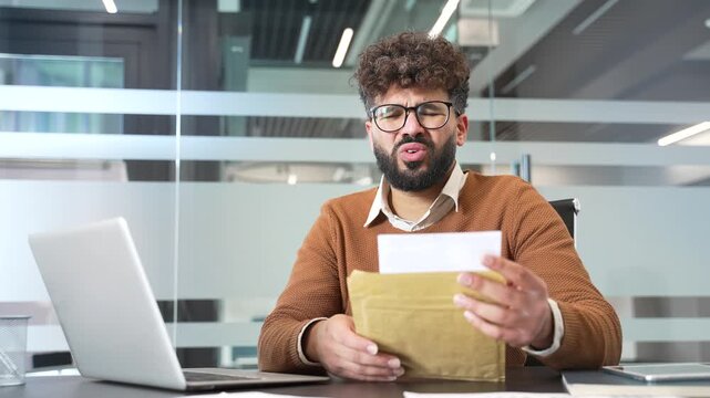 Upset disappointed businessman reading a letter with bad news sitting in business office. Worried male professional frustrated by receiving unpleasant negative notification after opening the envelope