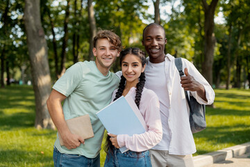 Fototapeta premium Three friends stand in a green park, smiling happily while holding books and folders. The sun shines brightly, creating a cheerful atmosphere for their gathering.