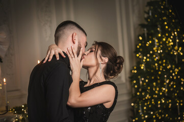 An elegant young couple in evening kiss near a Christmas tree in a studio during a candlelit photo...
