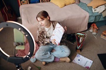 Portrait of Caucasian teenage blogger sitting on floor in bedroom holding certificate and looking...