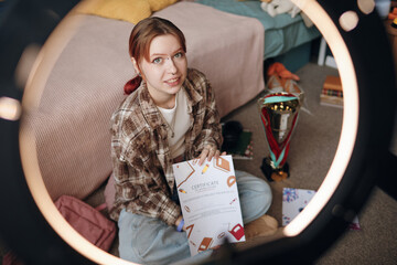 Portrait of Caucasian teenage girl sitting on floor in bedroom holding certificate and looking into...