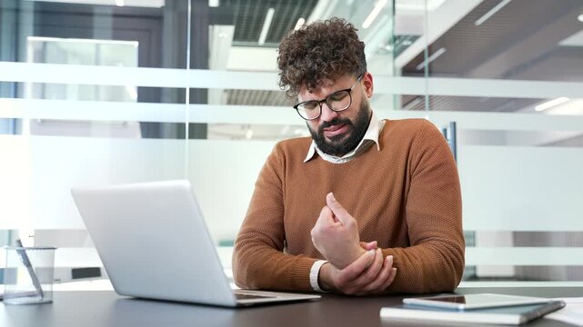 Unwell businessman suffering from wrist pain while working on laptop typing on keyboard while sitting at workplace in business office. Unwell man is massaging a joint, holds a sore spot with his hand