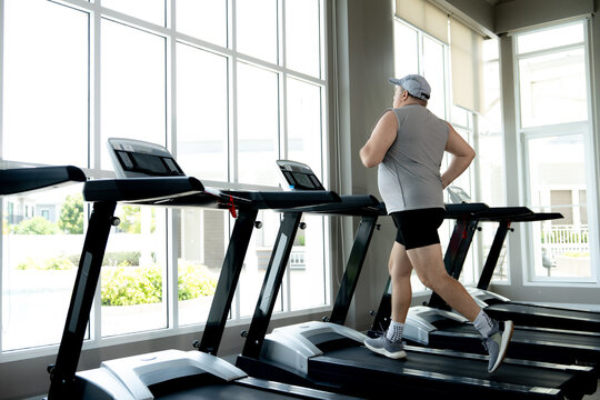 An active senior man is running on a treadmill at the fitness center. He enjoys his daily cardio workout indoors near a large window.
