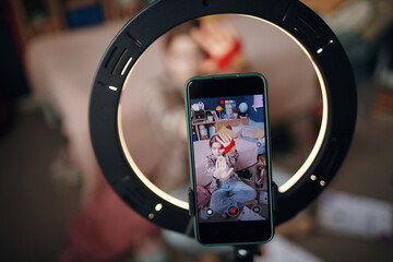 Caucasian teenage girl sitting on floor recording video with smartphone and ring light in bedroom, holding medal and gesturing toward camera