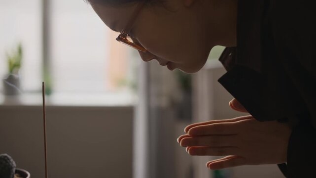Handheld shot of young Asian female employee bowing toward lit incense stick placed beside Buddha figure and fruit offering in office