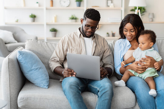 An African American family sits comfortably on a sofa at home. Parents engage with their baby boy on a laptop, watching cartoons and sharing moments with grandparents through a video call.
