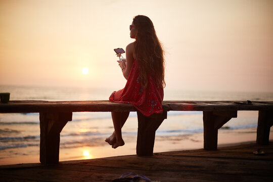 Young woman sitting on wooden bench holding cocktail glass gazing at ocean during sunset with long hair flowing down back enjoying tranquil seaside view