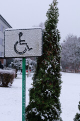 Wheelchair sign in snowfall. Wheelchair symbol sign stands beside snowy path and winter garden. Concept of accessibility awareness, urban communication, social messaging, and public design.