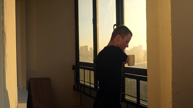 A man with a physical disability drinks coffee from a mug while looking out the window at sunrise on his balcony, enjoying a quiet morning moment