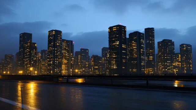 A cityscape with tall modem buildings, a clear sky with some clouds, and an empty road in the foreground. The buildings are illuminated, suggesting it is either dusk or early evening