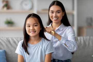 Fototapeta premium In a bright living room, a young mother gently combs her daughter's hair with a wooden brush. Both are relaxed on the sofa, sharing a tender moment filled with love and care.