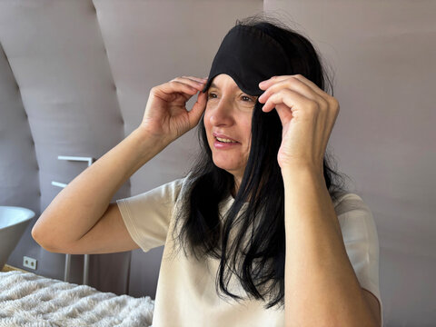 Adult brunette woman sitting in bed with a sleep mask, smiling and looking relaxed after a good night’s sleep, cozy morning routine, healthy sleep, wellness and self-care lifestyle