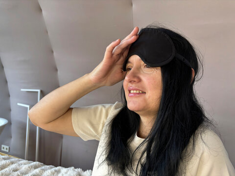 Adult brunette woman sitting in bed with a sleep mask, smiling and looking relaxed after a good night’s sleep, cozy morning routine, healthy sleep, wellness and self-care lifestyle