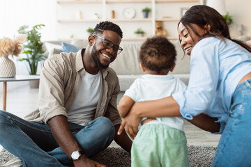 Obraz premium A happy black family sits on the carpet of their home, smiling while playing with their young son. The parents engage warmly with their toddler, celebrating joyful family moments.
