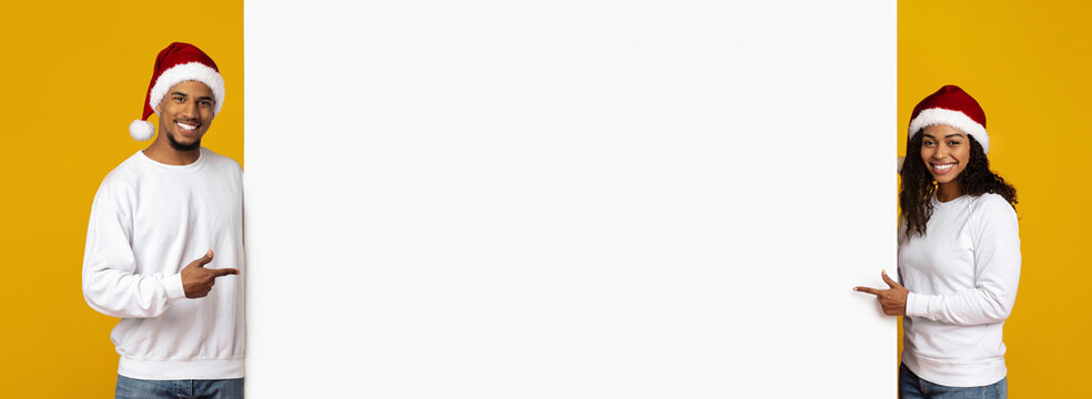 A cheerful couple wearing Santa hats stands on both sides of a large blank sign. They are smiling and pointing, ready for a festive celebration.