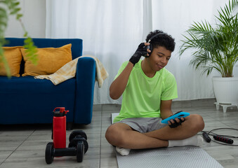 Brown skinned teenager exercising in his living room, sitting while using his phone and listening to music with headphones. Concept of home exercise and technology