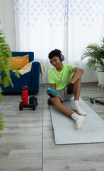 Brown skinned teenager working out at home, sitting on a mat entertained with his phone while listening to music. Concept of exercise and technology at home
