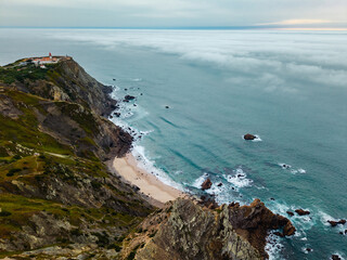 Aerial Coastal Landscape of Cabo da Roca Lighthouse, Steep Cliffs, and Turquoise Waters Meeting Foggy Atlantic Horizon in Sintra, Portugal