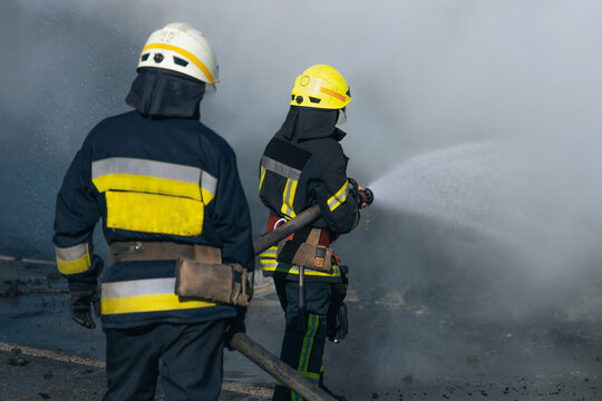 A close-up of two firefighters in protective uniforms and helmets. One of them directs a powerful stream of water or foam into an area of ​​dense smoke, working to extinguish the fire. - Powered by Adobe