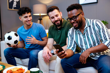 Three young adult men, two Caucasian and one Black, sitting on sofa watching TV together, one holding soccer ball, all smiling and interacting with smartphone during game