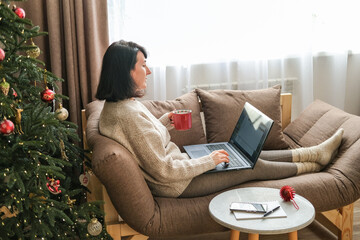 Middle-aged woman relaxing on sofa with laptop and red mug next to decorated Christmas tree and side table. Remote work, cozy holidays, online shopping, winter leisure