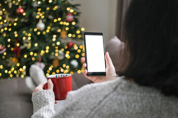 Woman in sweater holding red mug and smartphone with blank white screen with blurred Christmas tree and bokeh lights in background. Mobile app, cozy holidays, screen mockup, holiday shopping