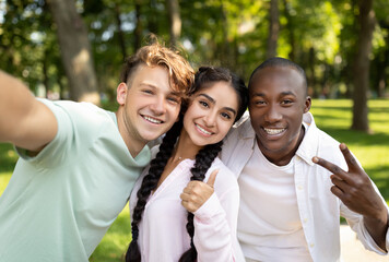 Three friends gather in a green park, smiling and taking a selfie. The sun shines brightly, creating a cheerful atmosphere as they pose with peace signs and thumbs up.