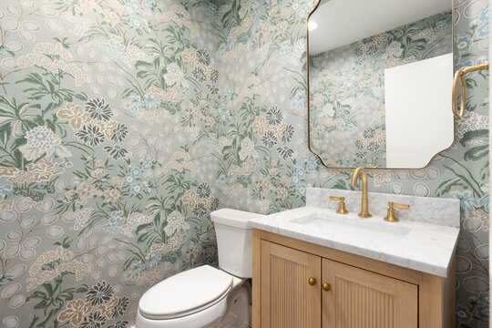 A half bathroom with a marble countertop on a white oak cabinet, gold faucet and mirror, and a green floral wallpaper.