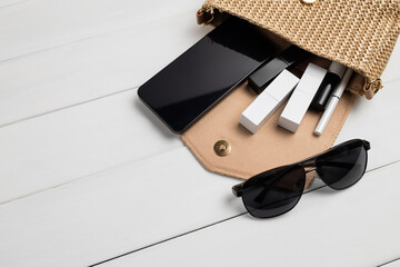 Open handbag with smartphone, cosmetics, makeup products and sunglasses on white wooden background with copy space, concept of beauty products and stylish accessories