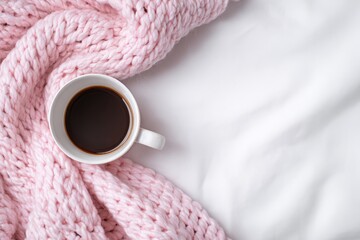 Cozy morning coffee on a white bed. A pink knitted blanket draped over a white bedsheet with a white coffee cup filled with dark coffee