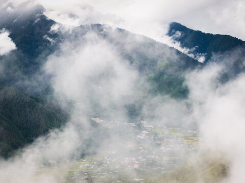 Aerial view of a valley shrouded in ethereal clouds, revealing glimpses of buildings nestled amidst the verdant landscape, Paro, Bhutan.