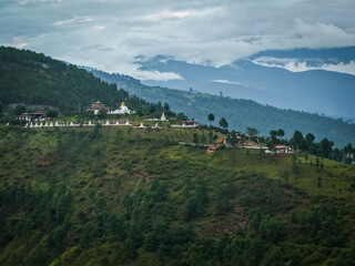 Aerial view of the white stupa dotting the verdant hillside near the temple complex, contrasting against the moody sky, Sopsokha, Punakha, Bhutan.