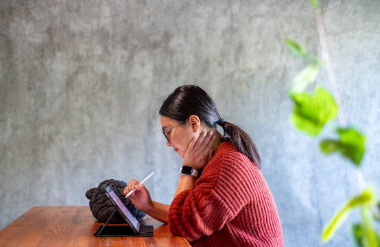 A woman working on a digital tablet with a stylus in a calm workspace. She focuses on creative tasks at a wooden desk, showing modern technology use and remote work lifestyle.