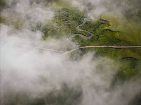Aerial view of the winding river snaking through the verdant Phobjikha Valley, a road cutting through the landscape, partially veiled by ethereal clouds, Nubding, Wangdue Phodrang, Bhutan.