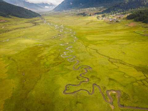 Aerial view of the serpentine river snaking through Phobjikha Valley's lush green expanse, bordered by dark forests and distant village, Nubding, Wangdue Phodrang, Bhutan.