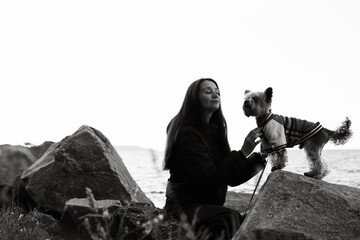 Black and white photo of a young woman with long hair gently touching her small Yorkshire terrier doggy in clothes on seaside rocks. Emotional portrait showing love connection of human and lovable pet