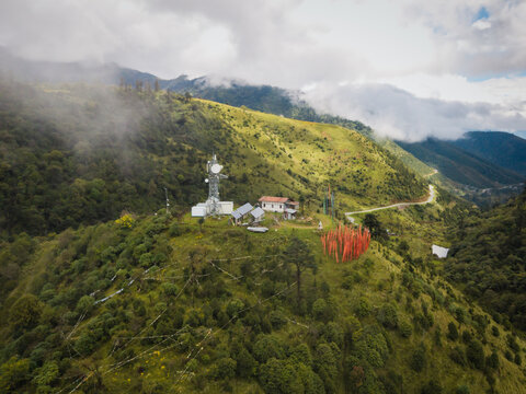 Aerial view of a telecom tower piercing the sky atop a lush, undulating mountain, where prayer flags dance vibrantly against the green slopes, Black Mountain range, Wangdue Phodrang, Bhutan.