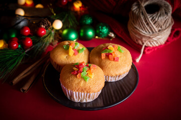 A warm and festive still life of Christmas-themed cupcakes topped with colorful candied fruit. The cupcakes are arranged with holiday decorations including pine branches, red berries, and rustic.