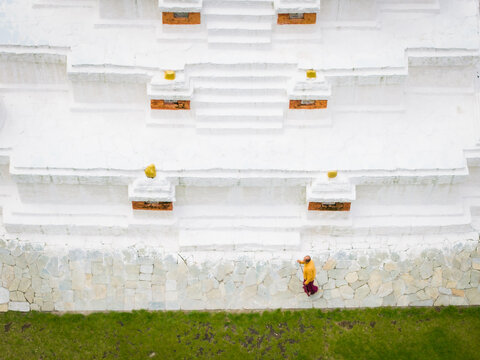 Aerial view of a lone figure in saffron robes walks along the stone wall of the Chendebji Chorten Stupa, its whitewashed tiers gleaming under the Bhutanese sun, Chendebji village, Trongsa, Bhutan.