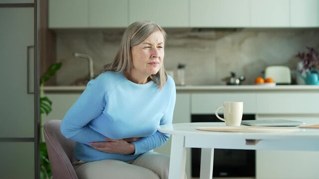 Senior female with gray hair feels stomach pain sitting at kitchen table at home. Sick elderly retired woman has heartburn, gastritis or poisoning. Suffers from spasms, constipation or bloating