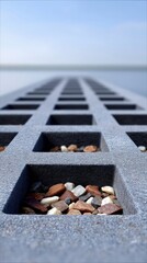 A close-up, low-angle view of a textured concrete grid filled with small stones, receding into a blurred background of calm water and a clear sky.
