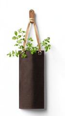 A dark brown fabric planter with green leaves is hanging from a wooden hook by leather straps against a clean white background, illuminated by soft natural ligh