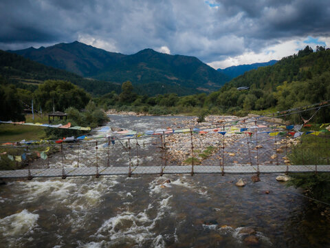 Aerial view of vibrant prayer flags fluttering across a rushing river, reflecting the dramatic sky above the verdant Tang Valley, Bumthang, Bhutan.