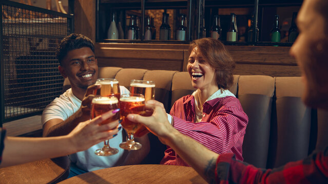 Friends clinking beer glasses at pub table during casual meetup. Concept of bar promotions, brewery advertising materials, nightlife marketing, social leisure, and beverage-focused branding.