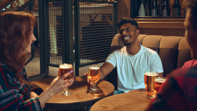 Friends chatting with beer glasses at wooden pub table. Concept of bar lifestyle content, brewery campaigns, social advertising, nightlife visuals, and relaxed drinking culture.
