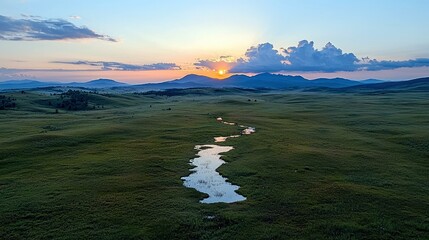 A winding stream flows through a vast, green meadow towards distant blue mountains under a vibrant sunrise sky with scattered clouds.