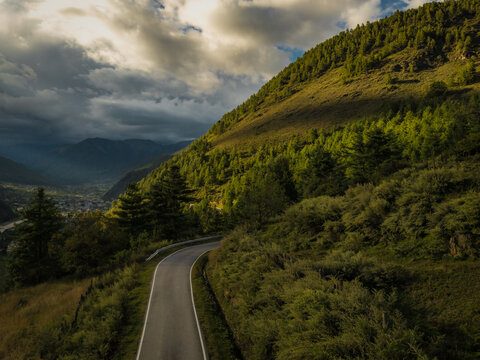 Aerial view of a winding road cutting through the verdant hillside, where light and shadow dance among the trees, leading towards the distant town, Paro, Paro, Bhutan.
